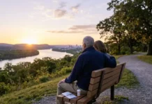 Living with CLL: Understanding Targeted Therapy for CLL and Its Impact Couple sitting on a wooden bench at sunset, overlooking a river valley with a distant city and a winding path nearby.