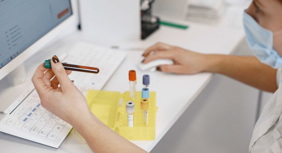 Lab technician wearing a mask holds a blood sample in a green-capped vial next to a yellow test-tube rack on a white lab bench with a computer keyboard nearby