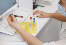 How Pharmaceutical Manufacturers Validate GMP Cleaning Protocols: ATP Bioluminescence Testing for Contamination Control Lab technician wearing a mask holds a blood sample in a green-capped vial next to a yellow test-tube rack on a white lab bench with a computer keyboard nearby
