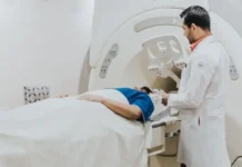 How AI-Powered Imaging Systems Are Solving Healthcare’s Radiologist Shortage Crisis Patient lying on an MRI table with head near the scanner, technician in a white coat preparing the machine.