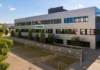 Syntegon opens new OSD Customer Center in Fellbach Exterior view of a white, multi‑story office building with many windows and a green SYNTEGON sign, trees lining the front, and a surrounding metal fence.