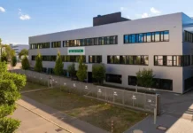 Syntegon opens new OSD Customer Center in Fellbach Exterior view of a white, multi‑story office building with many windows and a green SYNTEGON sign, trees lining the front, and a surrounding metal fence.